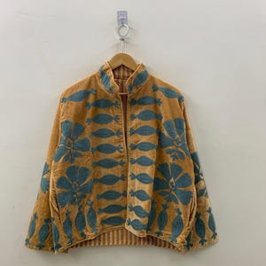 May include: A mustard-colored jacket with teal floral embroidery. The jacket has a mandarin collar and a striped lining. The embroidery features large flowers and leaf-like shapes. The jacket is hanging on a wooden hanger.