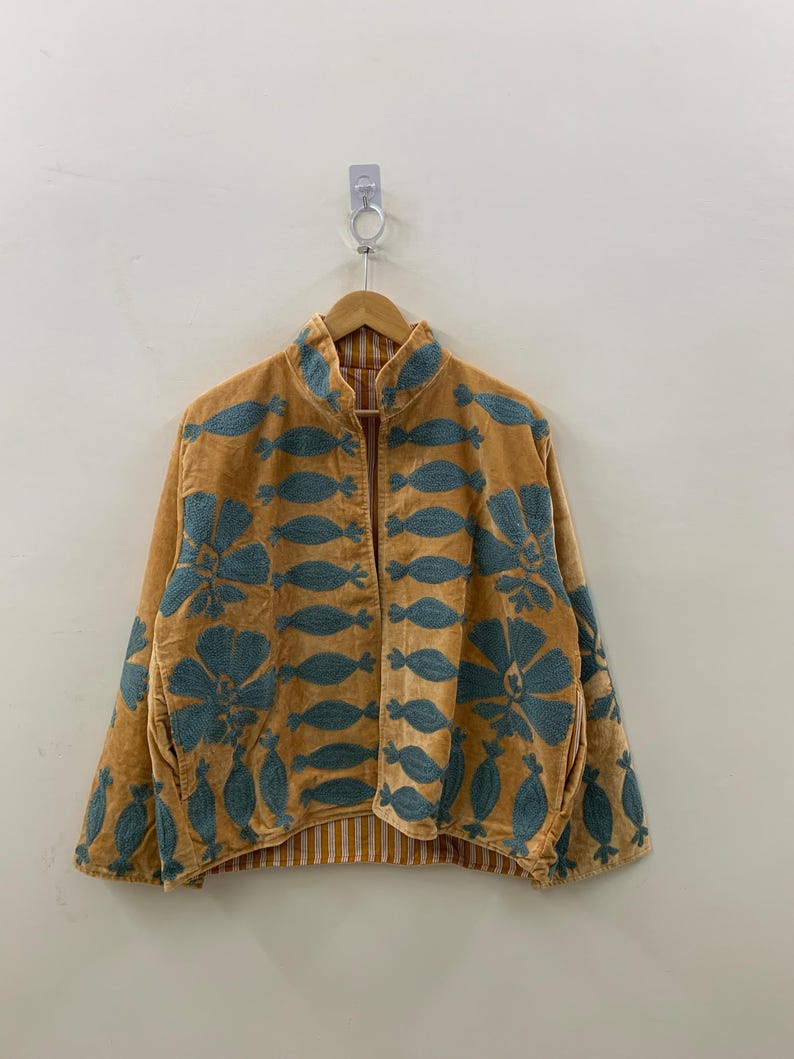 May include: A mustard-colored jacket with teal floral embroidery. The jacket has a mandarin collar and a striped lining. The embroidery features large flowers and leaf-like shapes. The jacket is hanging on a wooden hanger.