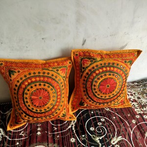 May include: Two square orange throw pillows with intricate embroidered circular patterns in red, green, and yellow. The pillows have a yellow border and are placed on a patterned fabric. The pillows are part of a home decor collection.