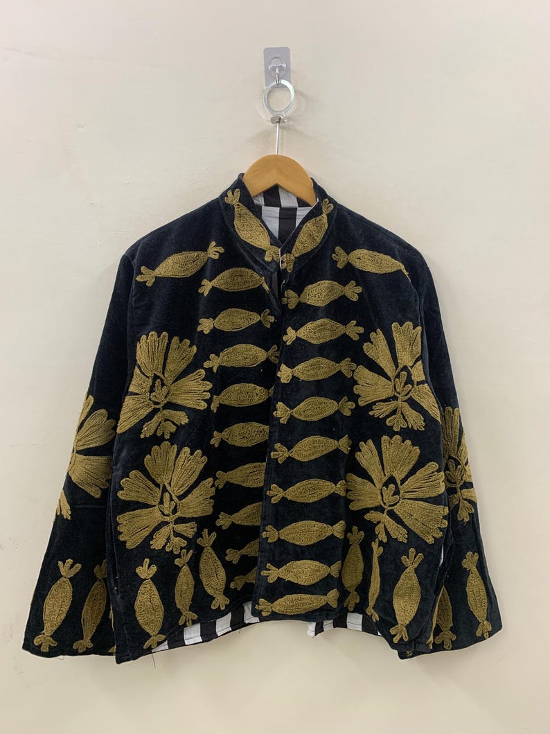May include: Black jacket with gold floral and abstract embroidery. The jacket has a high collar and a zipper closure. The lining is black and white checkered. The jacket is hanging on a wooden hanger.