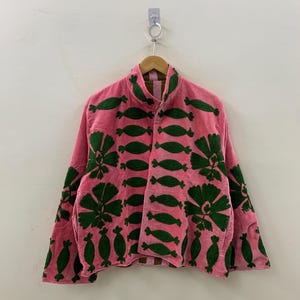 May include: A pink jacket with green embroidered floral and leaf patterns. The jacket is hanging on a wooden hanger against a white wall. The jacket has a high collar and long sleeves.