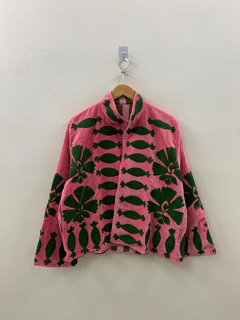May include: A pink jacket with green embroidered floral and leaf patterns. The jacket is hanging on a wooden hanger against a white wall. The jacket has a high collar and long sleeves.