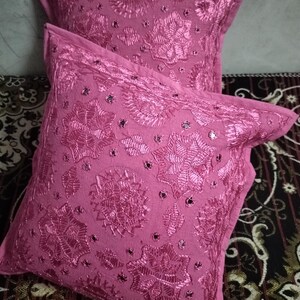 May include: Two pink decorative throw pillows with embroidered floral patterns and small mirrored accents. The pillows are square-shaped and rest on a patterned surface. The image includes the text "Shot on OnePlus World TextileExport".