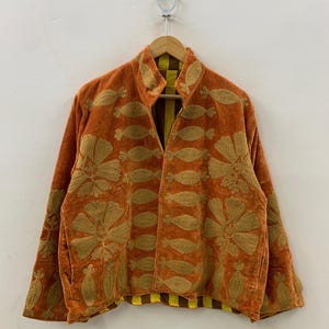 May include: An orange velvet jacket with gold floral embroidery. The jacket has a high collar and long sleeves. The interior lining is yellow and brown striped. The jacket is hanging on a wooden hanger.