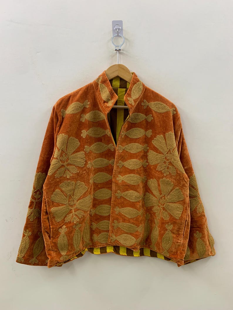 May include: An orange velvet jacket with gold floral embroidery. The jacket has a high collar and long sleeves. The interior lining is yellow and brown striped. The jacket is hanging on a wooden hanger.