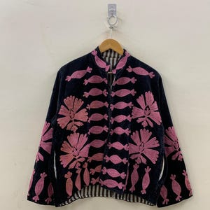 May include: A black jacket with pink floral embroidery and candy-shaped accents. The jacket has a high collar and a striped lining. The embroidery is detailed and covers the front and sleeves of the jacket. The jacket is hanging on a wooden hanger.