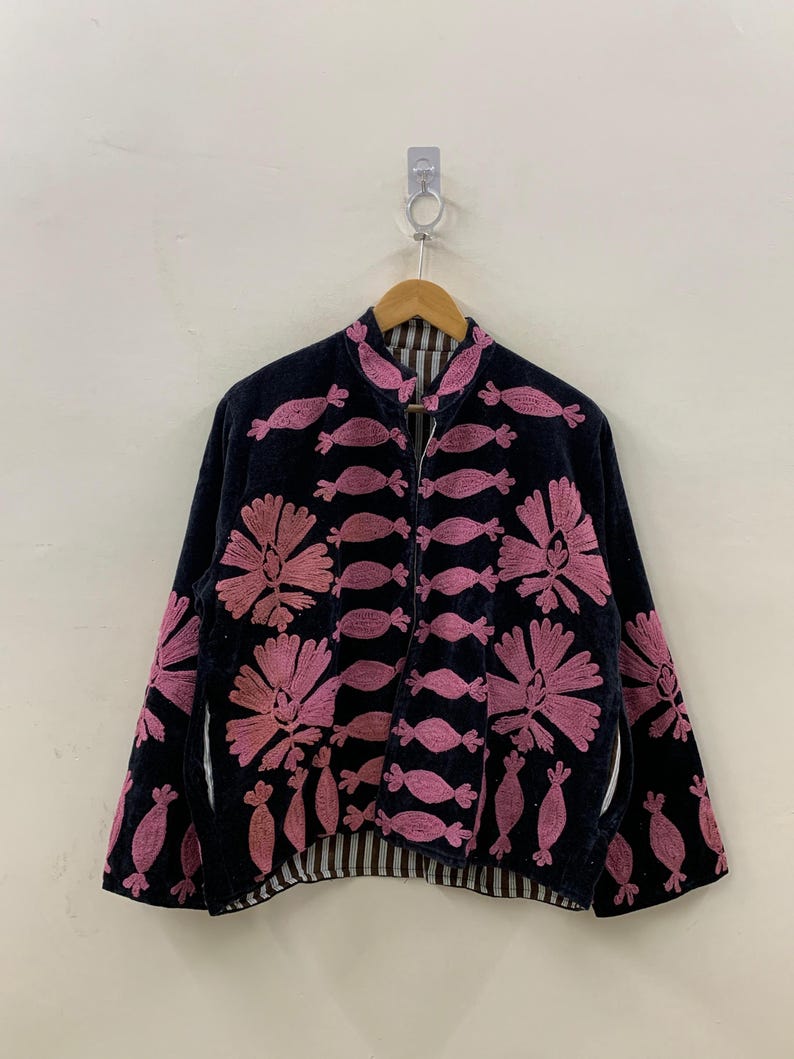 May include: A black jacket with pink floral embroidery and candy-shaped accents. The jacket has a high collar and a striped lining. The embroidery is detailed and covers the front and sleeves of the jacket. The jacket is hanging on a wooden hanger.