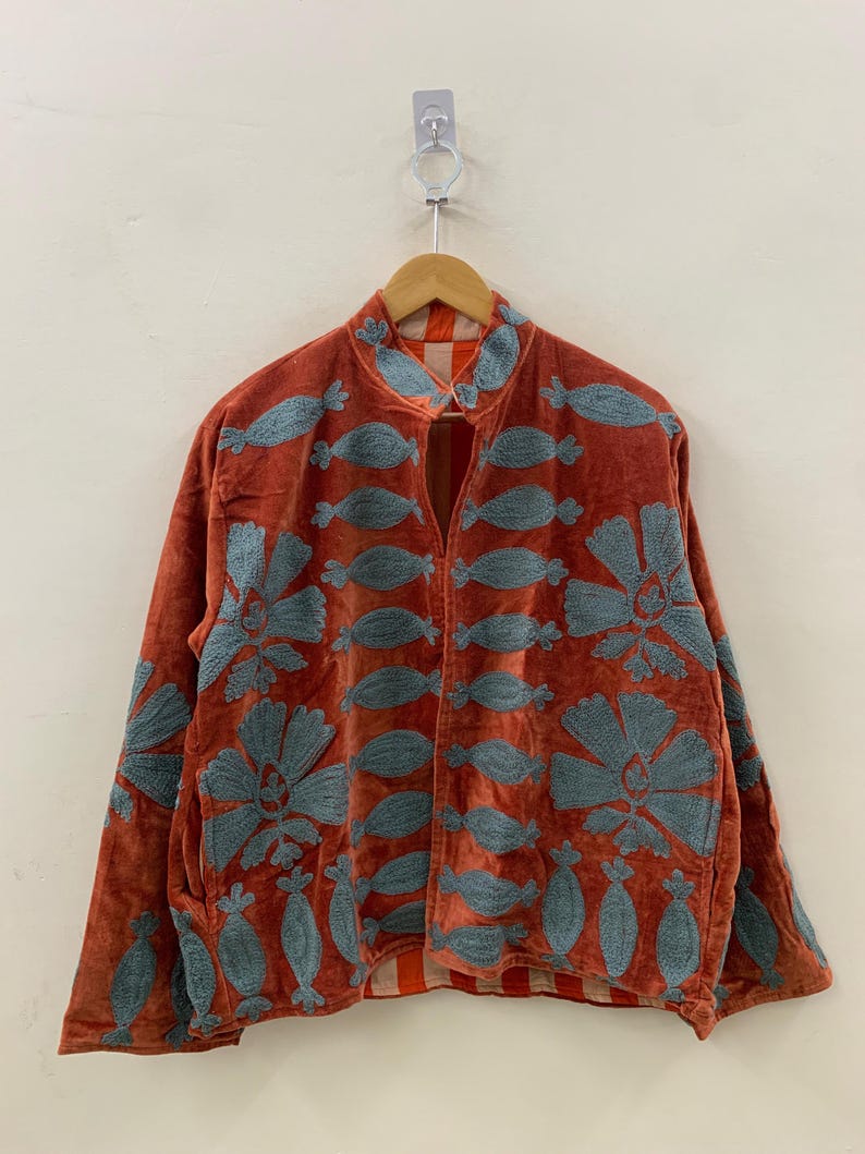 May include: Rust-colored velvet jacket with blue floral embroidery. The jacket features a mandarin collar and long sleeves. The embroidery includes large flowers and smaller, stylized leaf shapes. The jacket is hanging on a wooden hanger.
