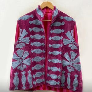 May include: A sleeveless, magenta-colored vest with a high collar, hanging on a wooden hanger. The vest is embroidered with light blue, stylized floral and candy-shaped designs. The interior lining is a bright pink color.