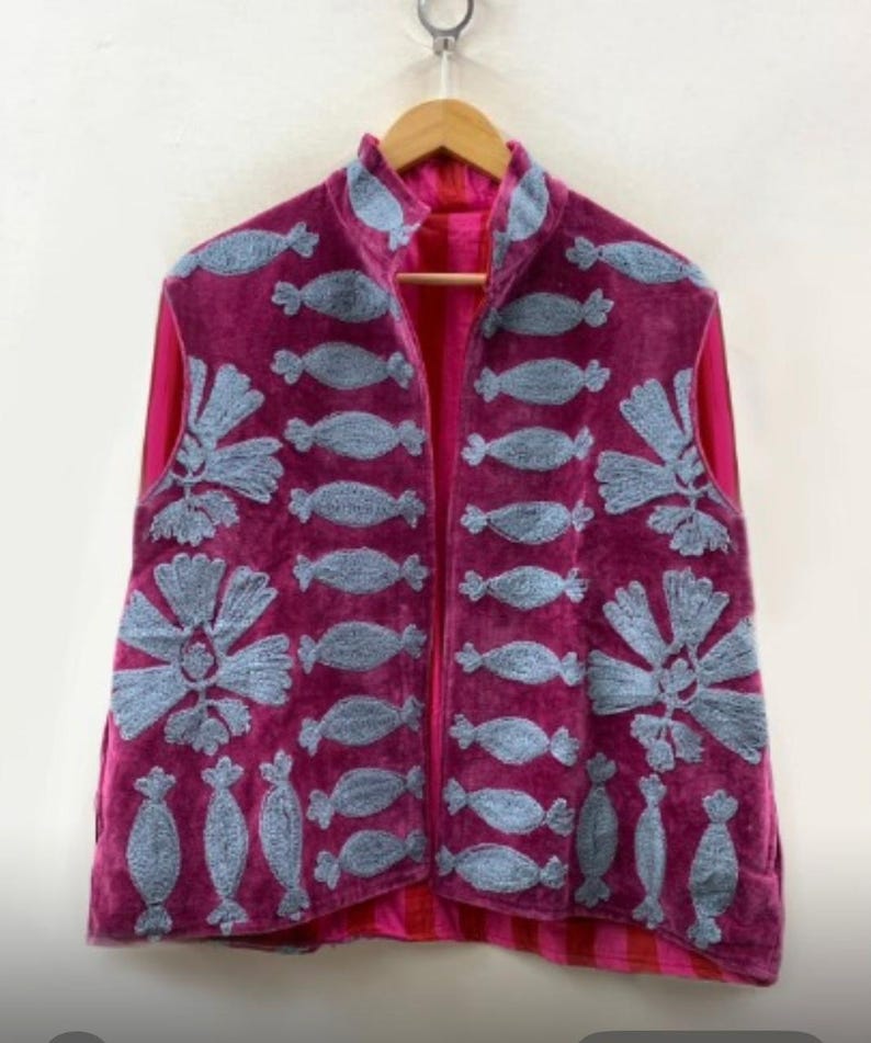 May include: A sleeveless, magenta-colored vest with a high collar, hanging on a wooden hanger. The vest is embroidered with light blue, stylized floral and candy-shaped designs. The interior lining is a bright pink color.