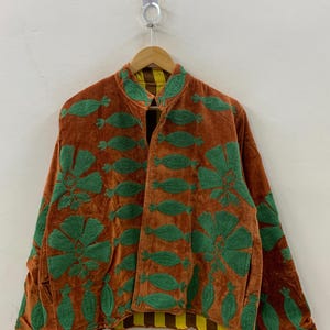 May include: A brown velvet jacket with green embroidered floral and leaf patterns. The jacket has a mandarin collar and a yellow and brown striped lining. The jacket is hanging on a wooden hanger.