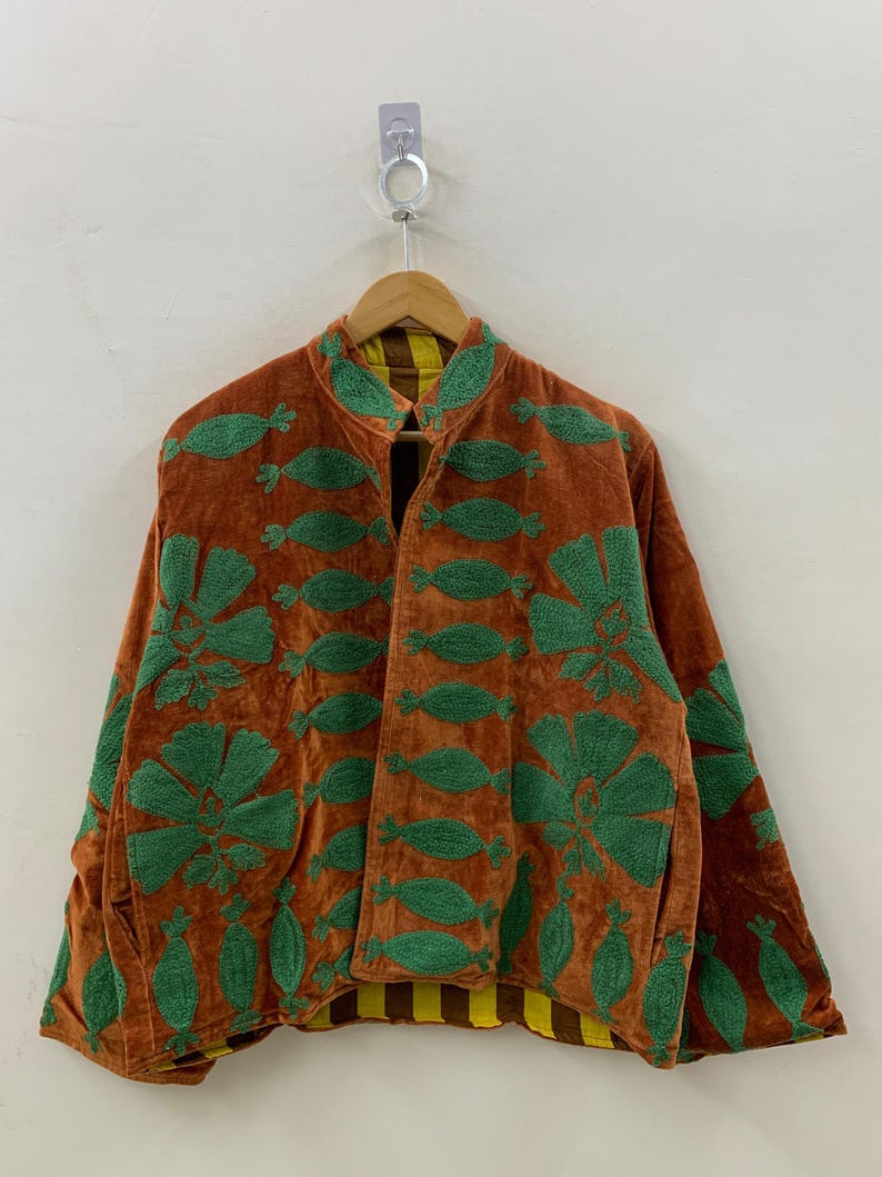 May include: A brown velvet jacket with green embroidered floral and leaf patterns. The jacket has a mandarin collar and a yellow and brown striped lining. The jacket is hanging on a wooden hanger.