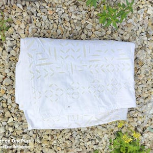 May include: White textile with a decorative pattern of geometric shapes and leaf-like designs. The fabric is folded and lies on a bed of small, light-colored stones. The text "Seller TextileExport" is visible.