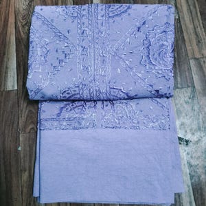 May include: A folded, light purple fabric set. The top piece features intricate white embroidery along the neckline and a detailed pattern. The bottom piece is a solid, lighter shade of purple. The set is displayed on a wooden surface.