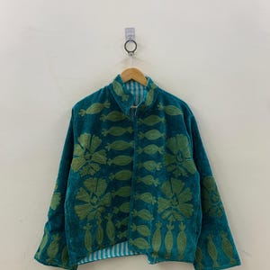 May include: Teal jacket with green floral embroidery. The jacket has a high collar and long sleeves. The embroidery is detailed and covers the front and sleeves. The jacket is hanging on a wooden hanger against a white wall.
