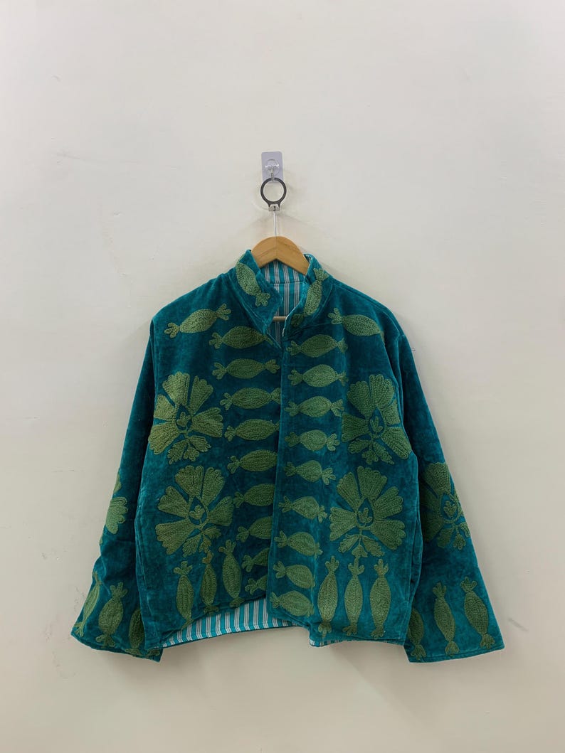 May include: Teal jacket with green floral embroidery. The jacket has a high collar and long sleeves. The embroidery is detailed and covers the front and sleeves. The jacket is hanging on a wooden hanger against a white wall.
