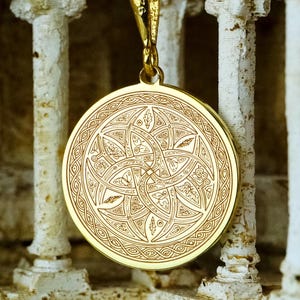Pode incluir: Um colar com pingente dourado. O pingente circular apresenta um design intrincado e simétrico de formas e linhas entrelaçadas. O pingente pende de uma delicada corrente dourada.
