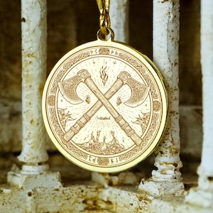May include: Gold-colored pendant necklace featuring a circular pendant with a detailed design of crossed axes and a landscape. The pendant hangs from a delicate gold chain. The background is a blurred architectural structure.