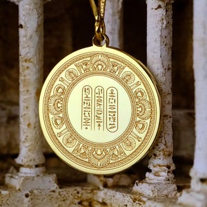 May include: A gold-colored pendant necklace with an engraved circular design and hieroglyphic text. The pendant hangs from a delicate gold chain. The background features a weathered, off-white architectural structure, creating a contrast with the shiny pendant.
