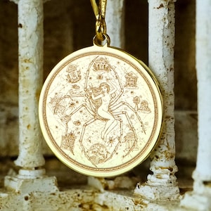 May include: A gold-colored pendant necklace featuring a circular charm with an engraved design. The charm depicts a figure with spider-like elements and other intricate details. The necklace is suspended from a delicate gold chain.