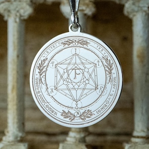 May include: A silver pendant necklace with a circular charm. The charm features an engraved geometric design with the letter "F" at the center, surrounded by text and laurel leaves. The pendant hangs from a delicate silver chain.