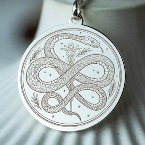 May include: A silver pendant necklace with a detailed engraved design of a snake coiled around a staff with a serpent. The circular pendant hangs from a delicate chain. The design is set against a blurred background.