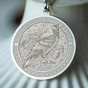 May include: A silver pendant featuring a detailed engraving of a raven. The round pendant has a decorative border and a bail for a chain. The raven is depicted with outstretched wings, set against a background of stars.