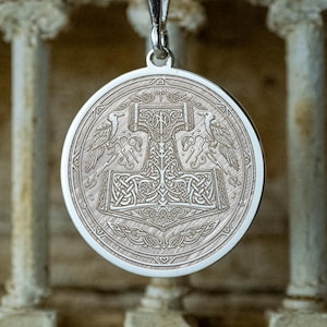 May include: A silver pendant necklace featuring a detailed circular pendant. The pendant showcases an engraved design of a hammer, possibly Mjolnir, with intricate patterns and avian figures. The necklace hangs from a delicate silver chain.