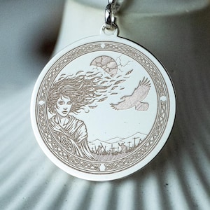 May include: A silver pendant necklace with a circular charm featuring an engraved design. The design includes a woman with flowing hair, a bird, a moon, and a landscape. The pendant hangs from a delicate silver chain.