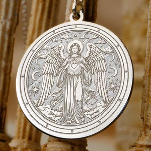 Puede incluir: Un collar con colgante de plata con un grabado detallado de un ángel con alas extendidas, un corazón y elementos celestiales. El colgante redondo cuelga de una delicada cadena. El fondo está borroso.