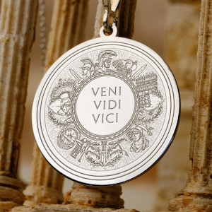 Puede incluir: Colgante plateado con un medallón redondo. El medallón presenta la frase latina «VENI VIDI VICI» en el centro, rodeada de grabados detallados de soldados romanos, armas y elementos arquitectónicos. El collar cuelga sobre un fondo de columnas antiguas.