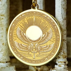 May include: A gold-coloured pendant with an Egyptian design. The pendant features a sun with wings, two birds, and an ankh symbol. The round pendant has detailed edges and hangs from a gold chain. The pendant's diameter is approximately 3 cm.