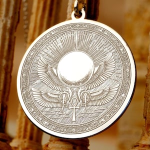 May include: A circular silver-toned pendant featuring an engraved design of a central sun disk with radiating rays, positioned above two symmetrical winged cobras flanking an ankh symbol, all enclosed within a decorative border of geometric patterns.