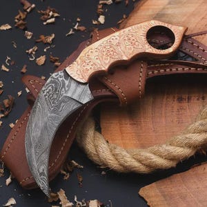 May include: A curved karambit knife with a patterned blade and a wooden handle featuring an engraved floral design. The knife rests in a brown leather sheath, with a rope and wood in the background.