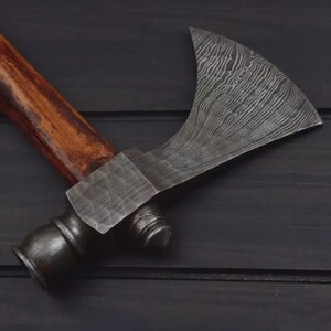 K&ouml;nnte beinhalten: Eine handgefertigte Axt mit einer gemusterten Stahlklinge und einem Holzgriff. Die Klinge hat eine gebogene Form und eine strukturierte Oberfl&auml;che. Der Griff ist aus dunklem Holz. Die Axt ist zum Hacken und Schneiden bestimmt.