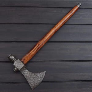 K&ouml;nnte beinhalten: Ein handgefertigtes Tomahawk mit Holzgriff und einer Stahlklinge mit Muster. Der Griff ist von tiefbrauner Farbe, und die Klinge hat ein strukturiertes, geschichtetes Aussehen. Das Tomahawk ist diagonal auf einer dunklen Holzoberfl&auml;che platziert.