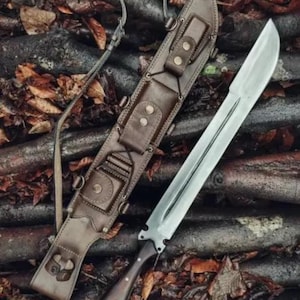 May include: A large, silver blade knife with a dark wooden handle and a brown leather sheath. The sheath has multiple compartments and is secured with straps and buttons. The knife and sheath are resting on a bed of leaves and wood.