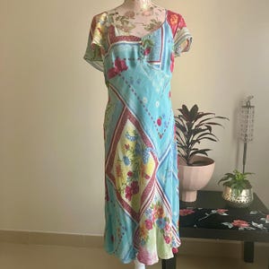 May include: A light blue dress with a floral pattern, featuring a square neckline and short sleeves. The dress showcases a mix of floral and geometric designs in red, yellow, and blue. The dress is displayed on a mannequin.