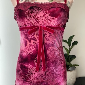 May include: A burgundy velvet camisole with a paisley pattern and lace trim. The camisole features a velvet bow at the waist and thin velvet straps. The garment is displayed on a mannequin. The overall theme is vintage lingerie.