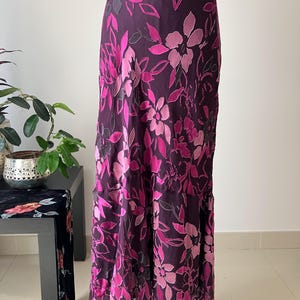 May include: A long, flowing skirt with a dark purple background and a floral pattern in shades of pink and fuchsia. The skirt has a high waist and a slight flare at the bottom, creating an elegant silhouette.