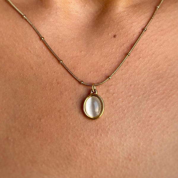 May include: A gold-colored necklace with a delicate chain and a small, oval pendant. The pendant features a white, opalescent stone set in a gold frame. The necklace is shown against a skin background.