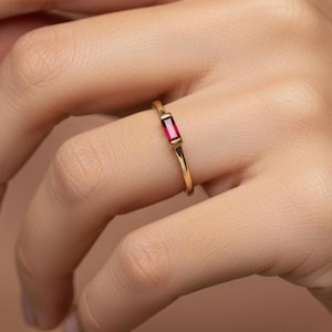 Puede incluir: Un anillo de oro con una piedra preciosa rectangular de color rubí. El anillo es sencillo y elegante, con una banda fina y una sola piedra vibrante. El anillo se muestra en un dedo.