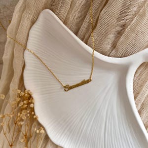 May include: A delicate gold-colored necklace with the name "Samantha" in cursive script. The necklace rests on a white, leaf-shaped dish, with a neutral-toned fabric in the background. The chain is thin and the pendant is in a stylish font.
