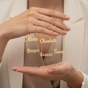 May include: Gold-colored name necklaces spelling out Olivia, Charlotte, Harper, Isabella, and Emma. The necklaces are displayed against a neutral background, showcasing the delicate chains and personalized pendants.