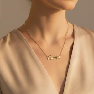 May include: A gold-colored necklace with the name "Emma" in a cursive font. The necklace is worn against a beige-colored top. The chain is delicate, and the pendant is centered on the chest. The overall aesthetic is elegant and minimalist.