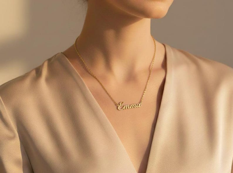 May include: A gold-colored necklace with the name "Emma" in a cursive font. The necklace is worn against a beige-colored top. The chain is delicate, and the pendant is centered on the chest. The overall aesthetic is elegant and minimalist.