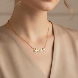 May include: A gold-colored necklace with the name "Amanda" in cursive script. The pendant hangs from a delicate chain. The necklace is worn against a beige top.