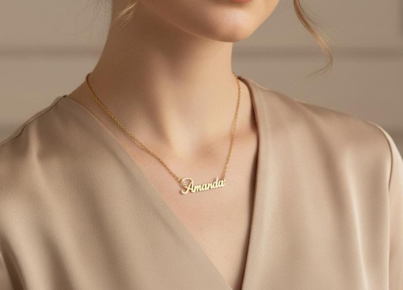 May include: A gold-colored necklace with the name "Amanda" in cursive script. The pendant hangs from a delicate chain. The necklace is worn against a beige top.