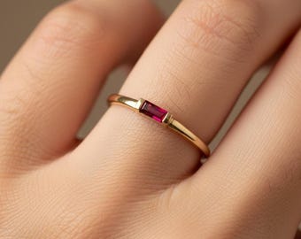 May include: A gold ring with a rectangular, deep red gemstone set in the center. The ring is simple and elegant, with a thin band and a minimalist design. The gemstone is the focal point, adding a pop of color.