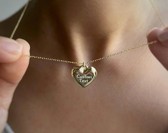 May include: A gold-colored heart-shaped pendant necklace with a delicate chain. The pendant has the words "Custom Text" engraved on it. The necklace is being held up, showcasing the pendant.